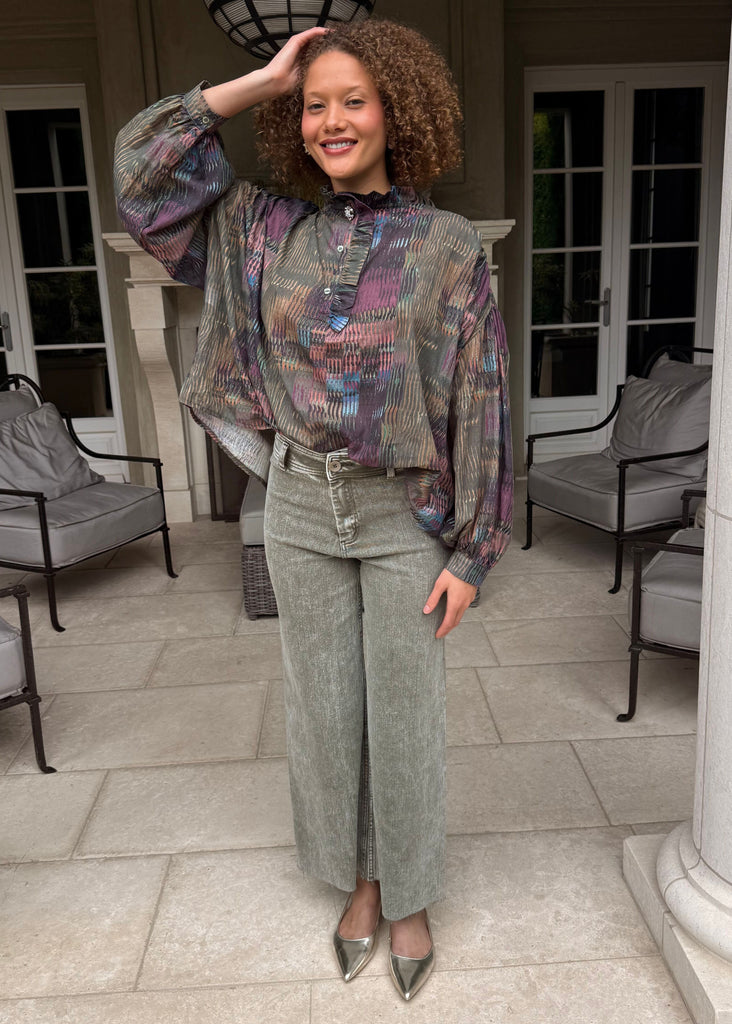 Woman posing in a patterned top and green pants on a patio with columns and chairs.