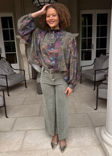 Woman posing in a patterned top and green pants on a patio with columns and chairs.