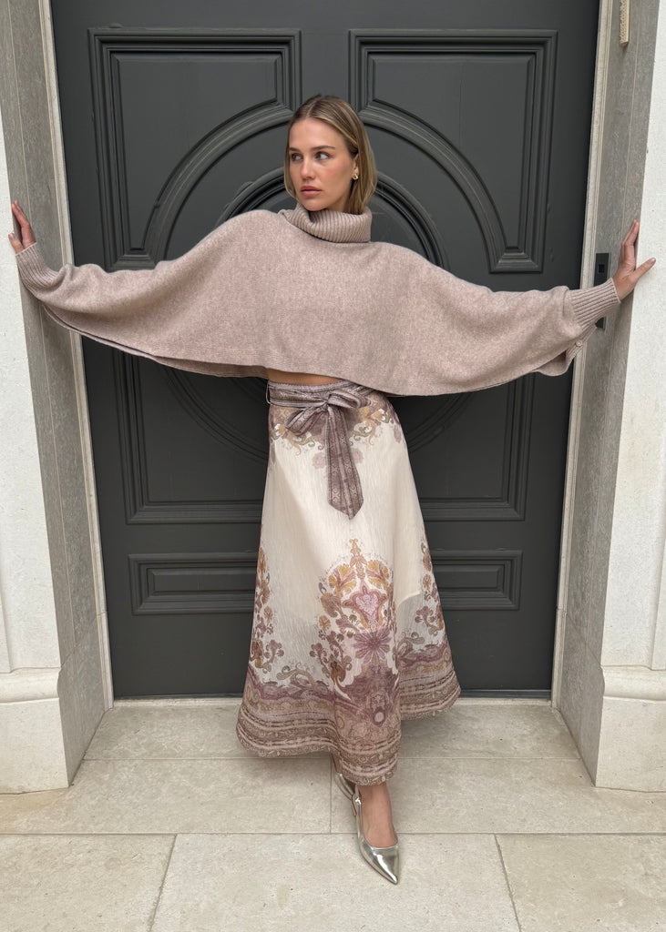 Woman wearing a beige cape and floral dress standing in front of a dark door.