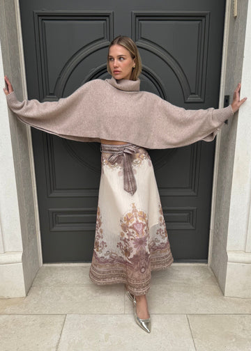 Woman wearing a beige cape and floral dress standing in front of a dark door.