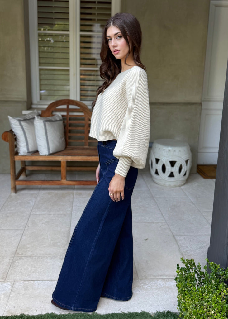 Woman in a white top and blue pants standing in a patio area with a bench and pillows.