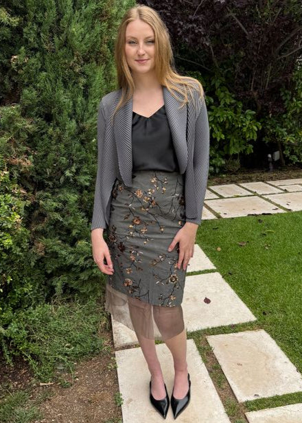 Woman in a gray blazer and skirt standing on a garden path with greenery.