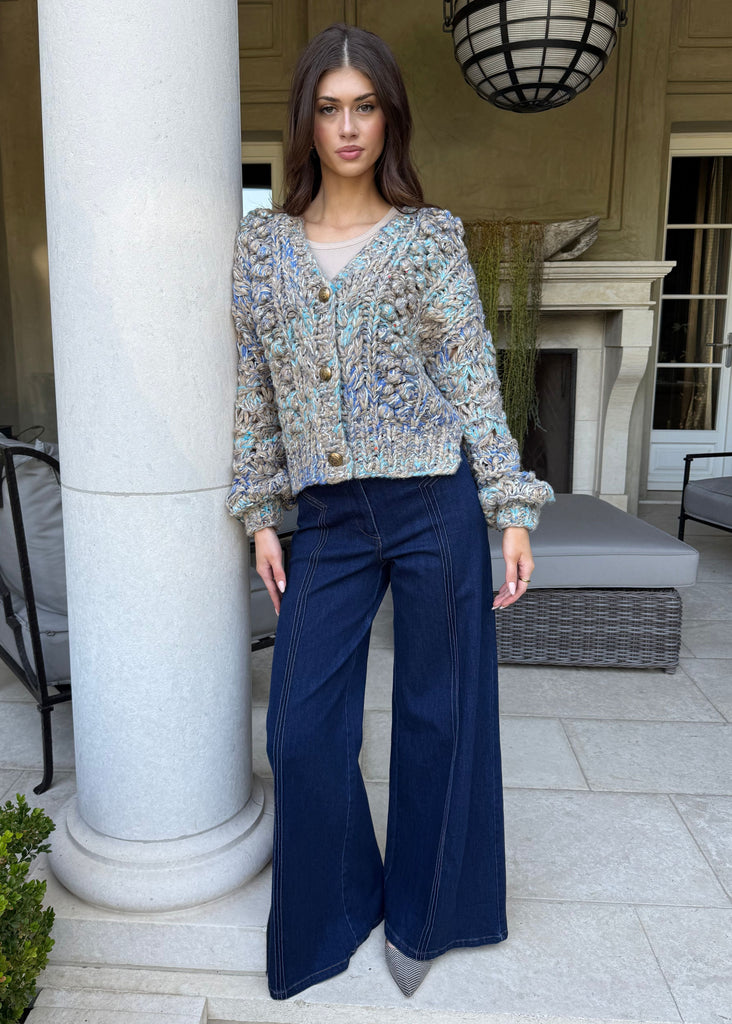 Woman in a patterned blouse and blue pants standing in front of a building entrance.