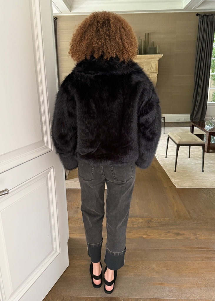 Person wearing a black fur coat in a room with wooden flooring and a window.