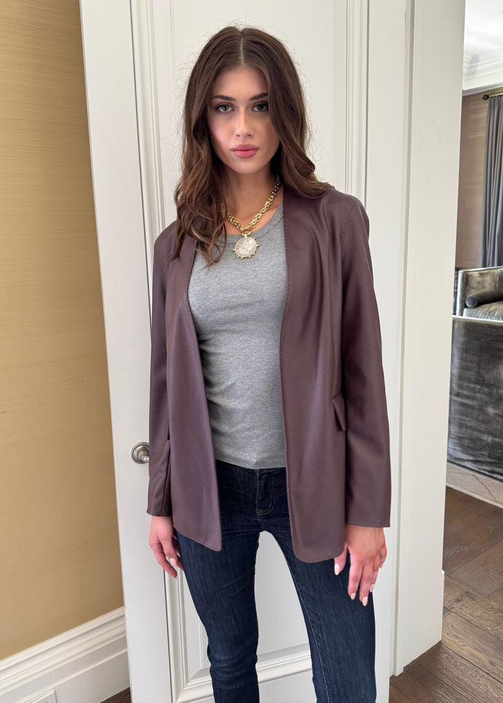 Woman wearing a brown blazer over a gray top and blue jeans, standing in a room with a mirror.