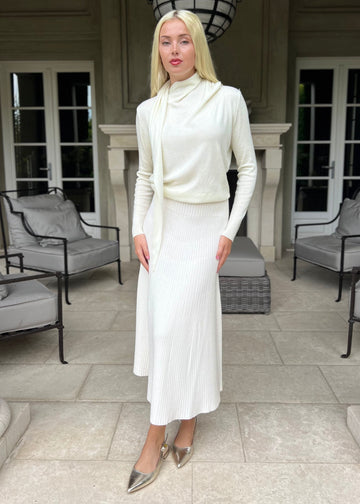 Woman in a white outfit standing in a stylish outdoor setting with chairs and a fireplace.