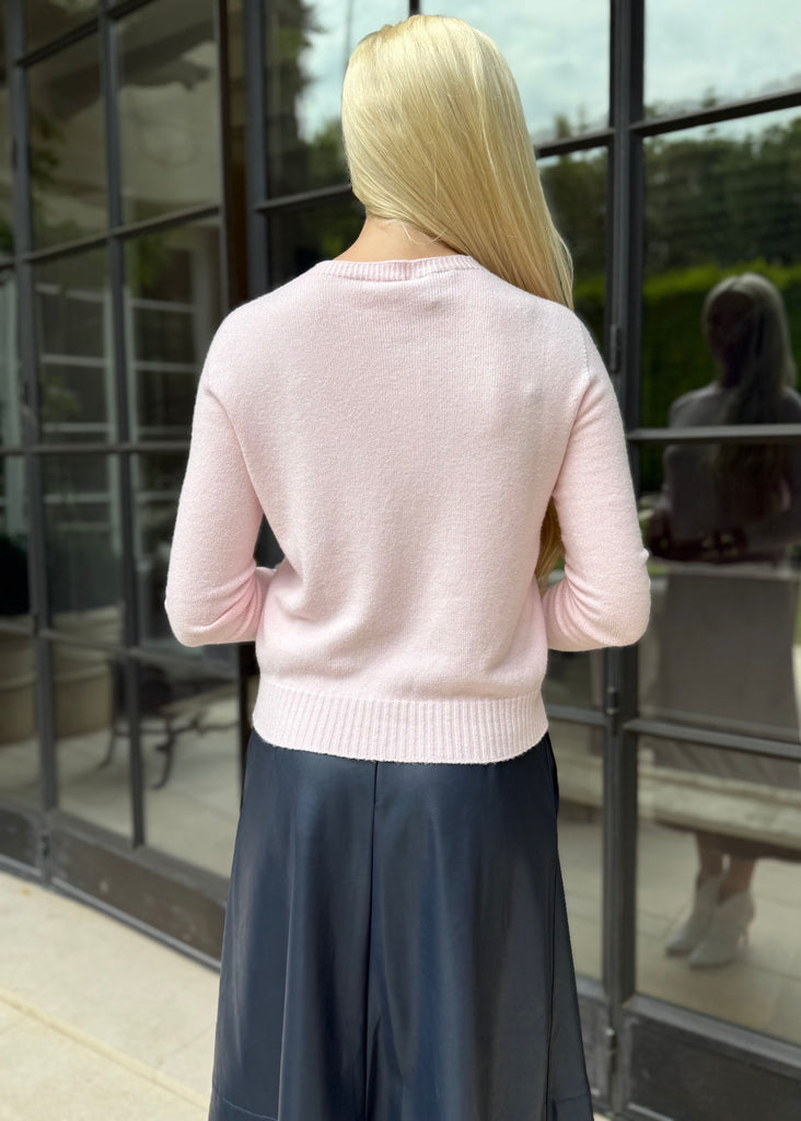 Woman wearing a pink sweater and dark pants standing in front of a glass door.