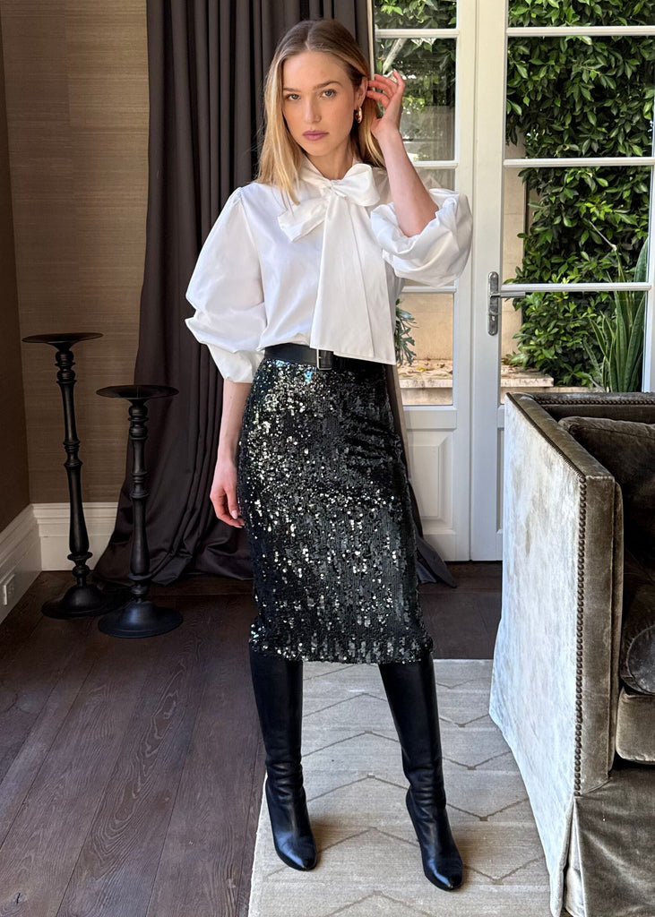 Woman in a stylish outfit standing in a living room with a couch and large windows.