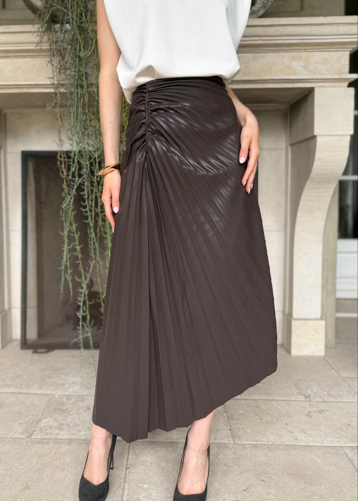 Person wearing a white top and brown pleated skirt standing in front of a building entrance.