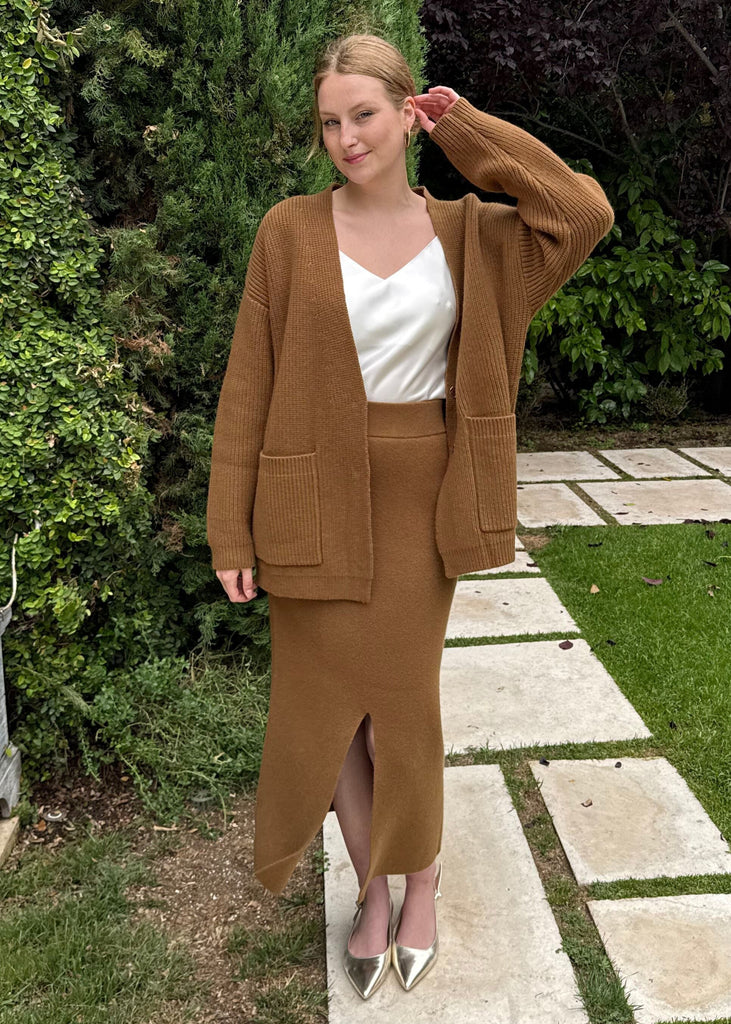 Woman in a brown suit standing in a garden with greenery and stone pathways.