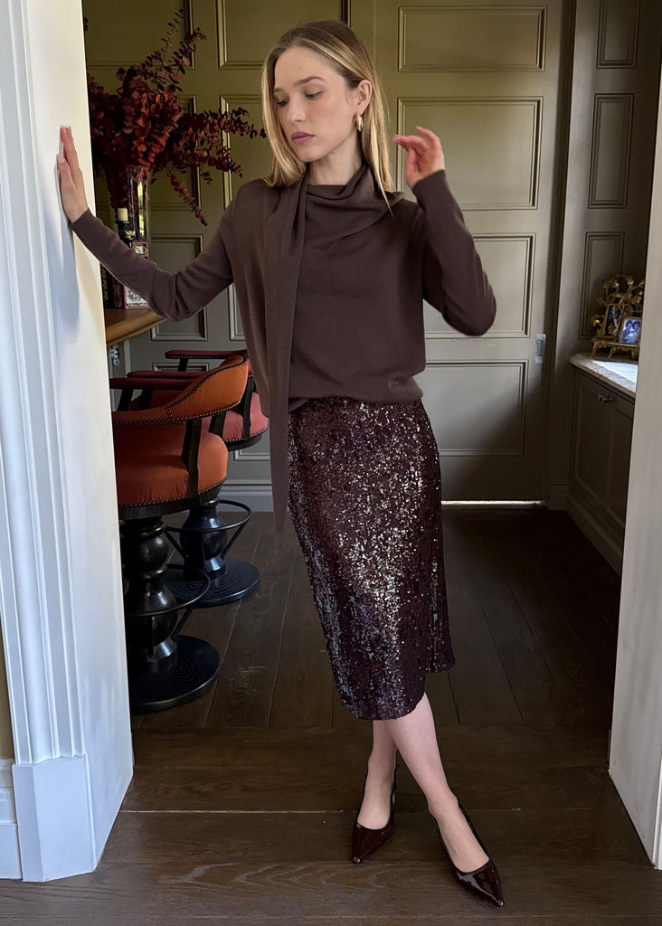 Woman in a brown top and sequin skirt standing in a room with a mirror.