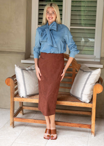 Woman in a blue shirt and brown skirt standing on a wooden bench with cushions.