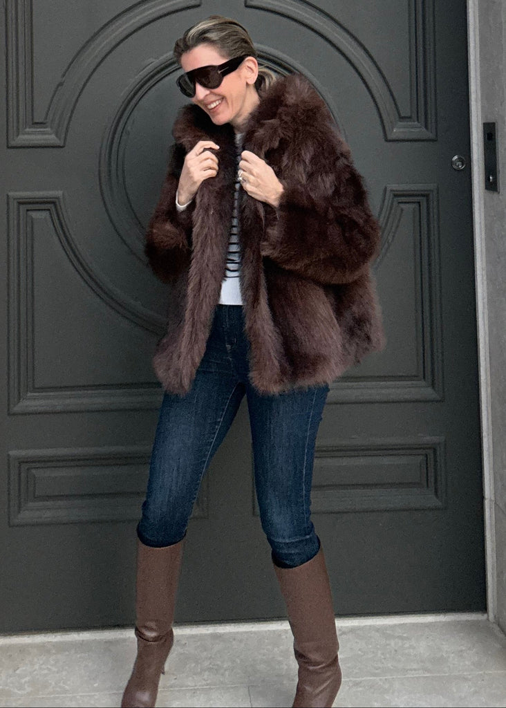 Person taking a mirror selfie wearing a fur coat in front of a decorative door.