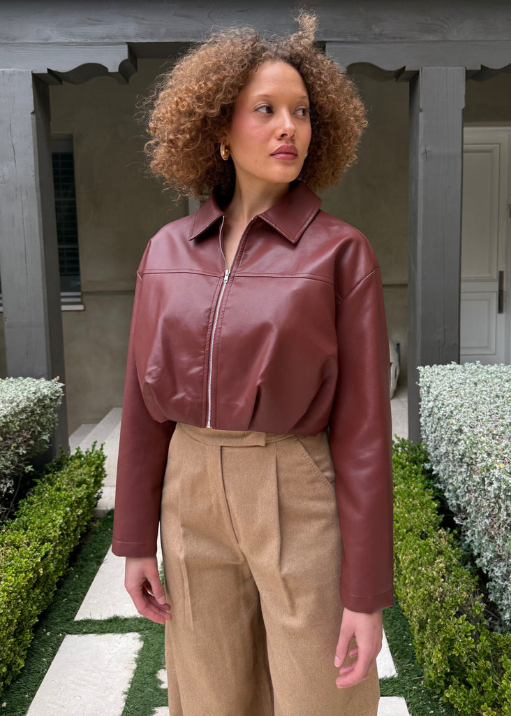 Woman wearing a maroon leather jacket and beige pants standing in front of a building with greenery.