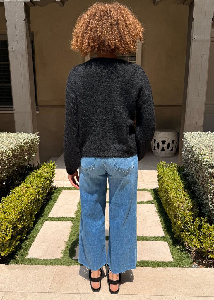 Person wearing a black sweater and blue jeans standing in a garden with hedges and a building in the background.