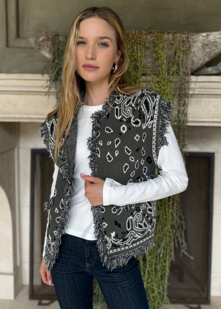Woman wearing a patterned vest over a white shirt in front of a fireplace.
