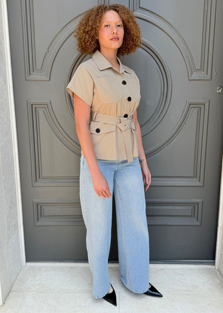 Person wearing a beige shirt with a belt and light blue jeans standing in front of a gray door.
