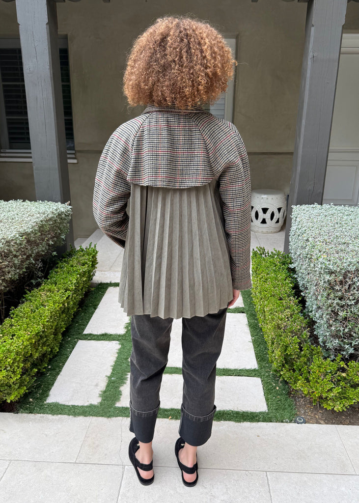 Person wearing a plaid jacket and gray pants standing in a garden with manicured hedges.