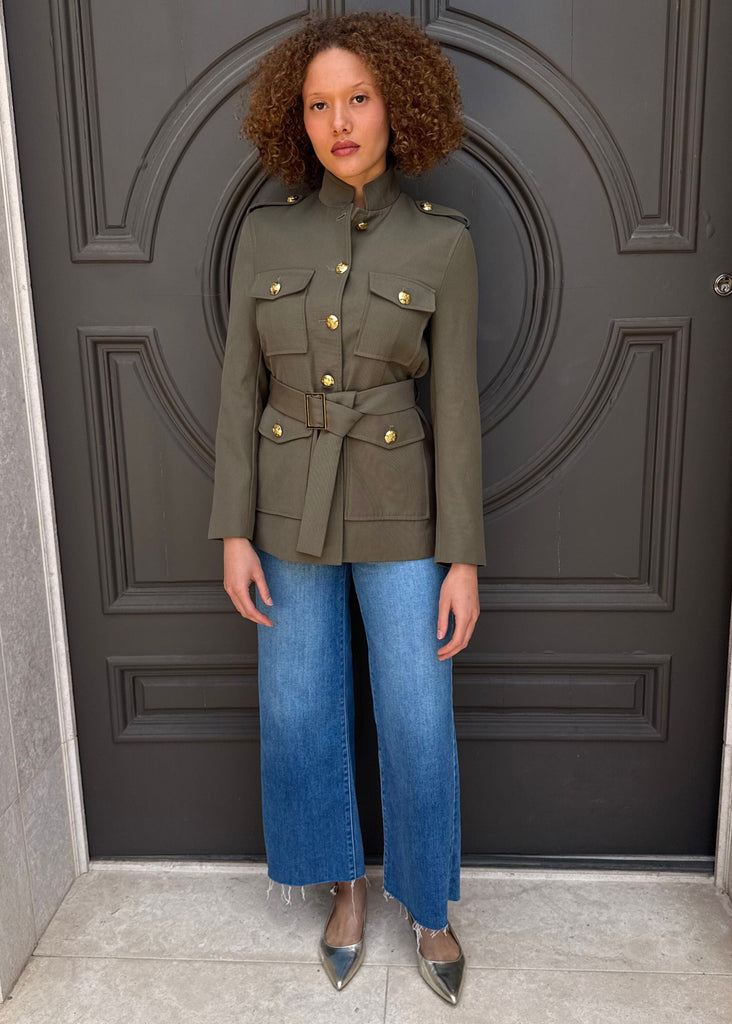 Woman wearing a green military-style jacket and blue jeans standing in front of a dark gray door.
