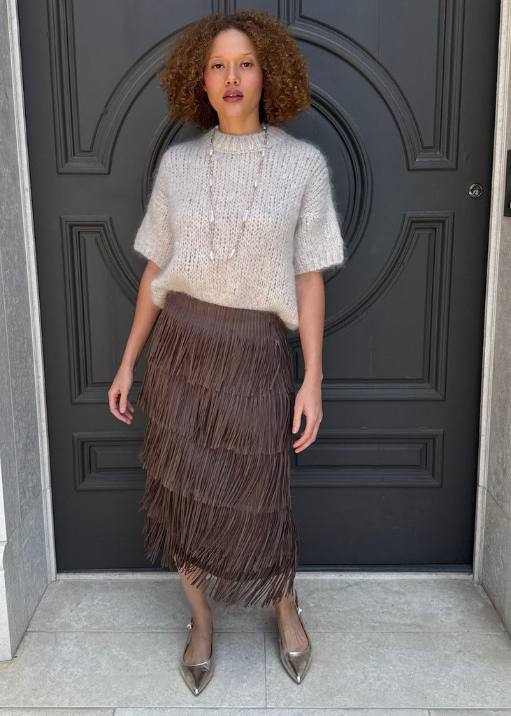 Woman standing in front of a dark gray door wearing a white top and brown fringed skirt.