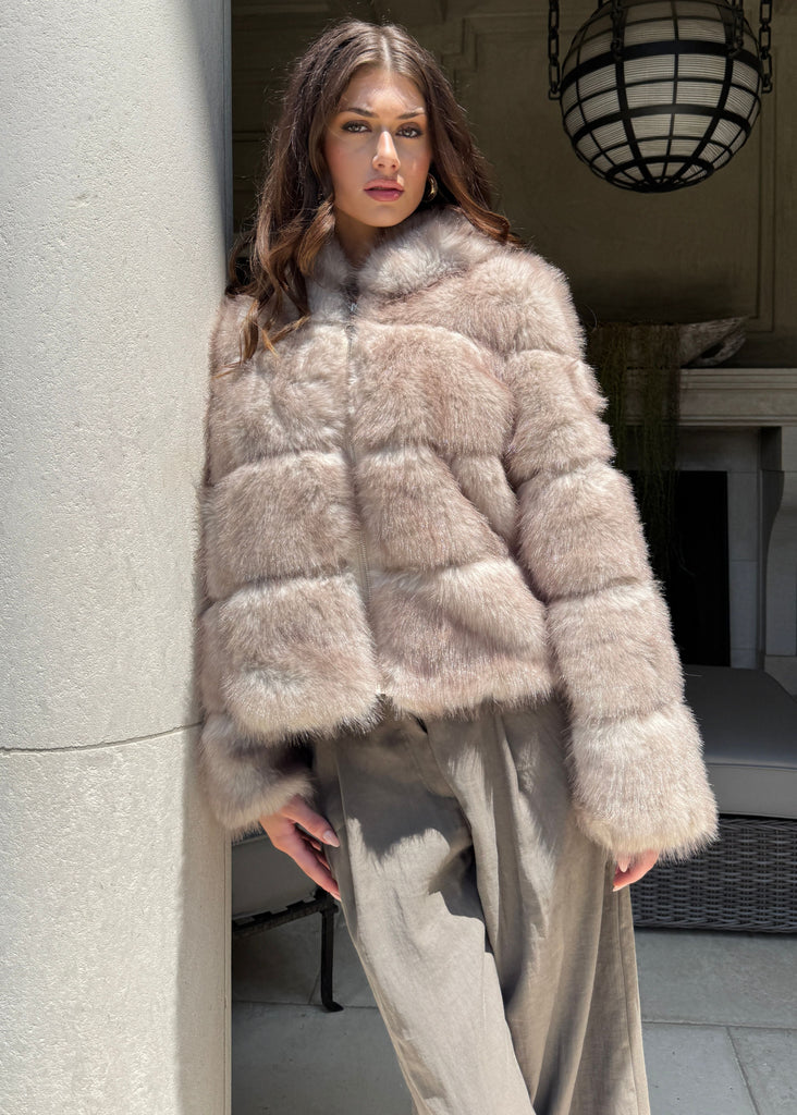 Woman wearing a beige fur coat standing in a room with a fireplace and large light fixture.