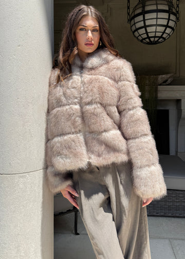 Woman wearing a beige fur coat standing in a room with a fireplace and large light fixture.