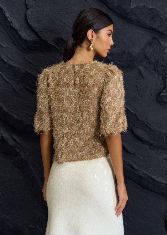 Woman wearing a textured brown top and white skirt against a dark stone wall.