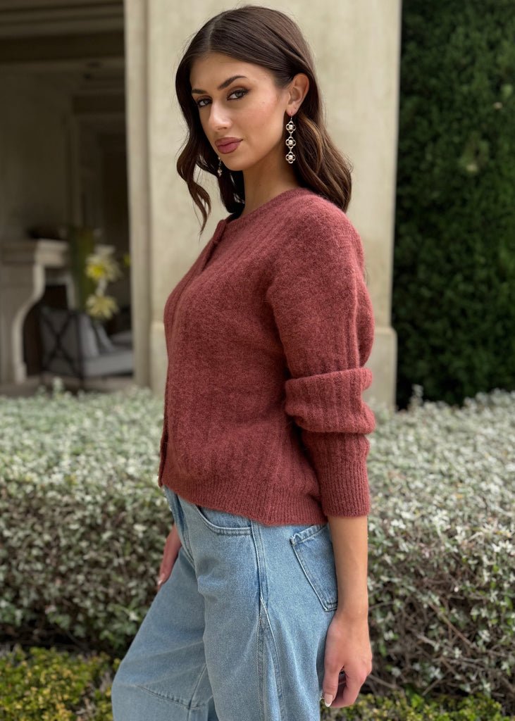 Woman wearing a maroon sweater and light blue jeans standing outdoors.
