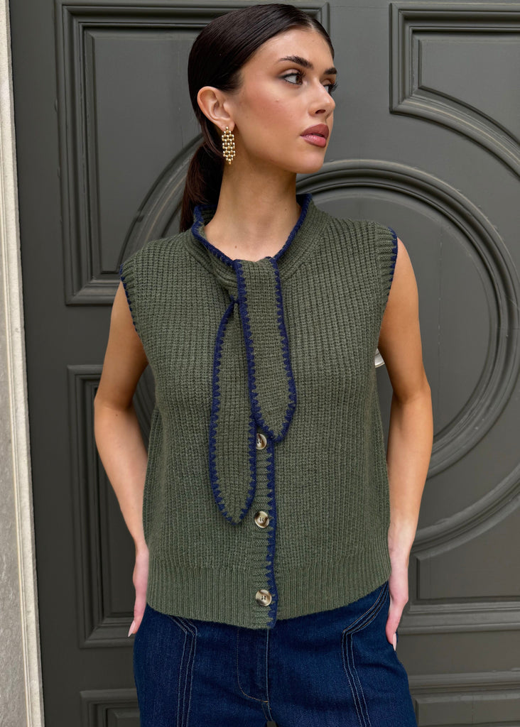 Woman wearing a green sleeveless knitted top with blue trim against a gray door.