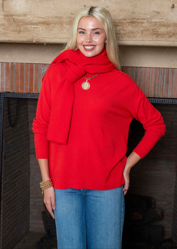 Woman wearing a red sweater and scarf standing in front of a fireplace.