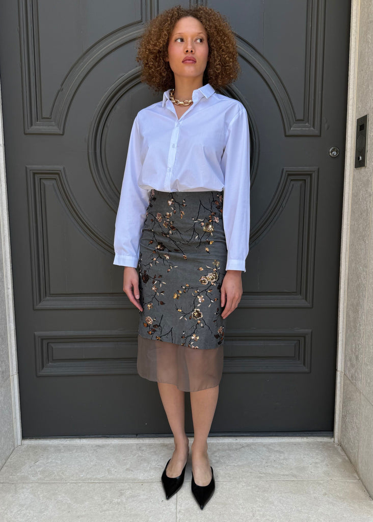 Woman standing in front of a dark gray door wearing a light blue shirt and patterned skirt.