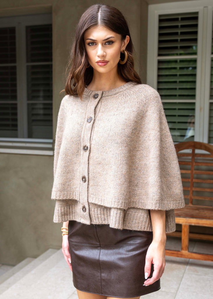 Woman wearing a beige cape over a brown leather skirt outdoors.