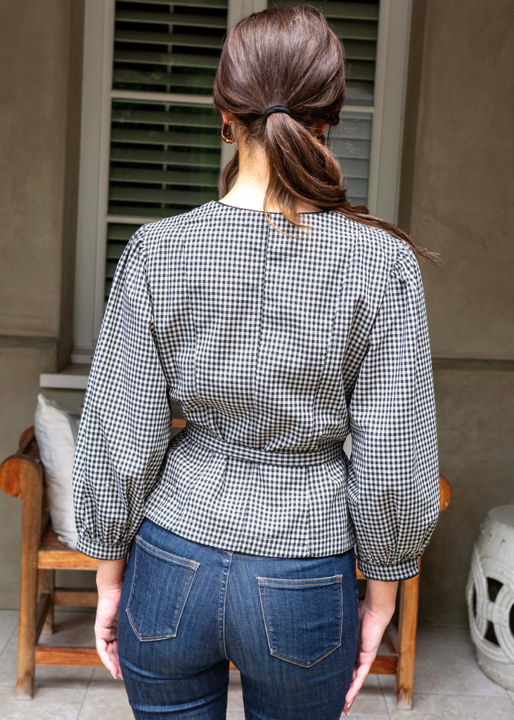 Person wearing a checkered shirt and jeans standing in front of a window with shutters.
