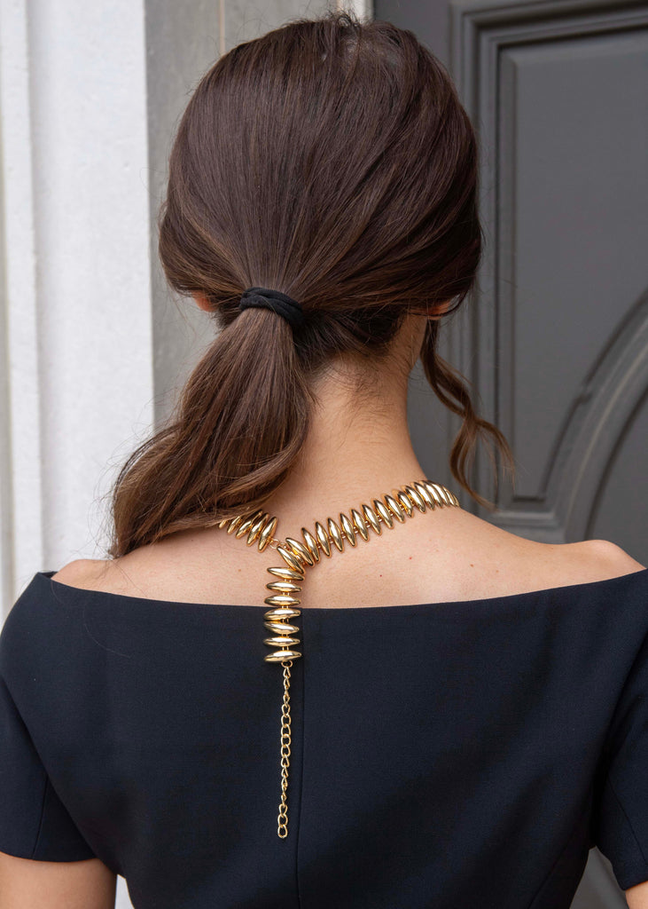 Woman with a stylish hairdo and gold necklace, standing against a neutral background