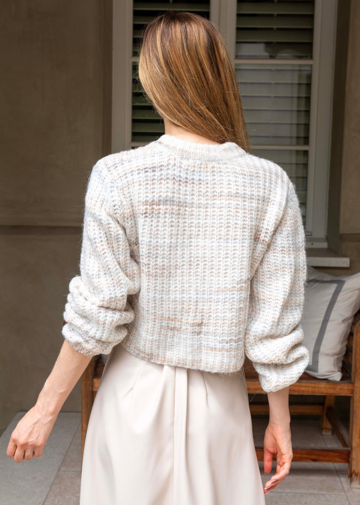 Woman wearing a textured white sweater and light-colored pants standing in front of a window with shutters.