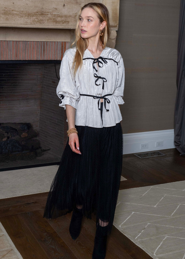 Woman wearing a white blouse with black lace details and a black skirt in a room with a fireplace.