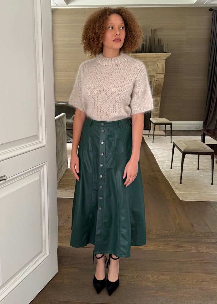 Woman standing in a room wearing a beige top and green skirt.