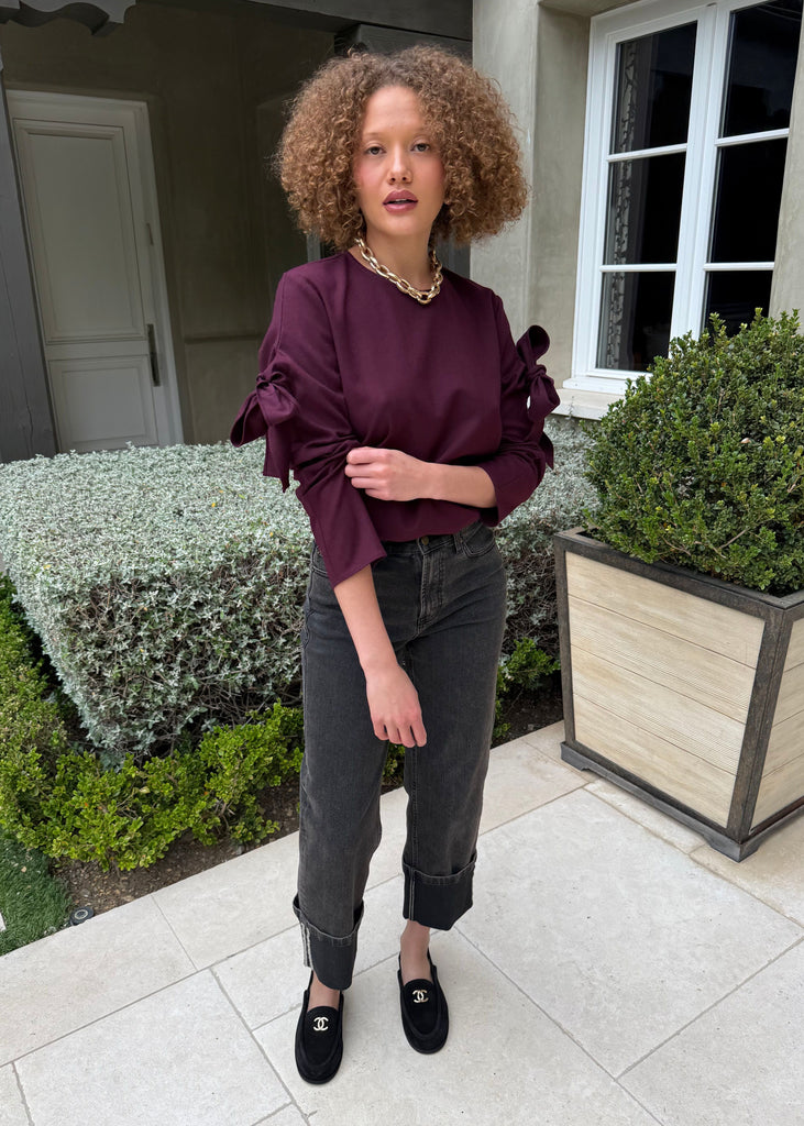 Woman in a burgundy blouse and gray pants standing outdoors near a building.