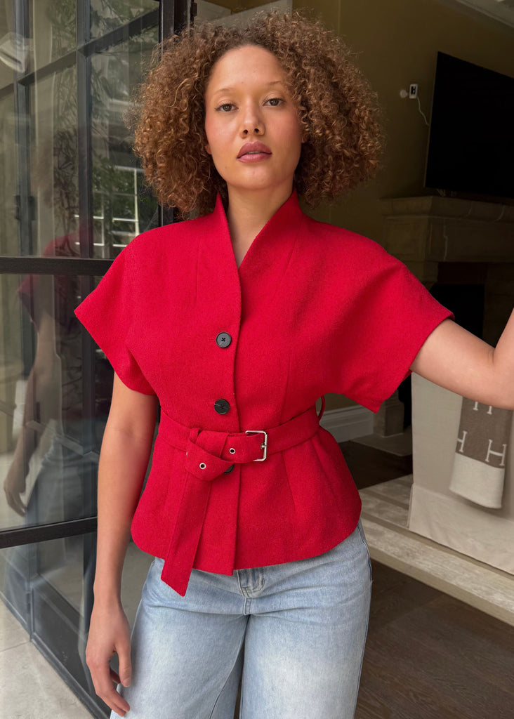 Woman wearing a red blouse with a belt and light blue jeans, standing in a doorway.