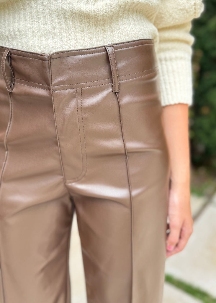 Brown leather pants worn with a light-colored sweater against a blurred green background