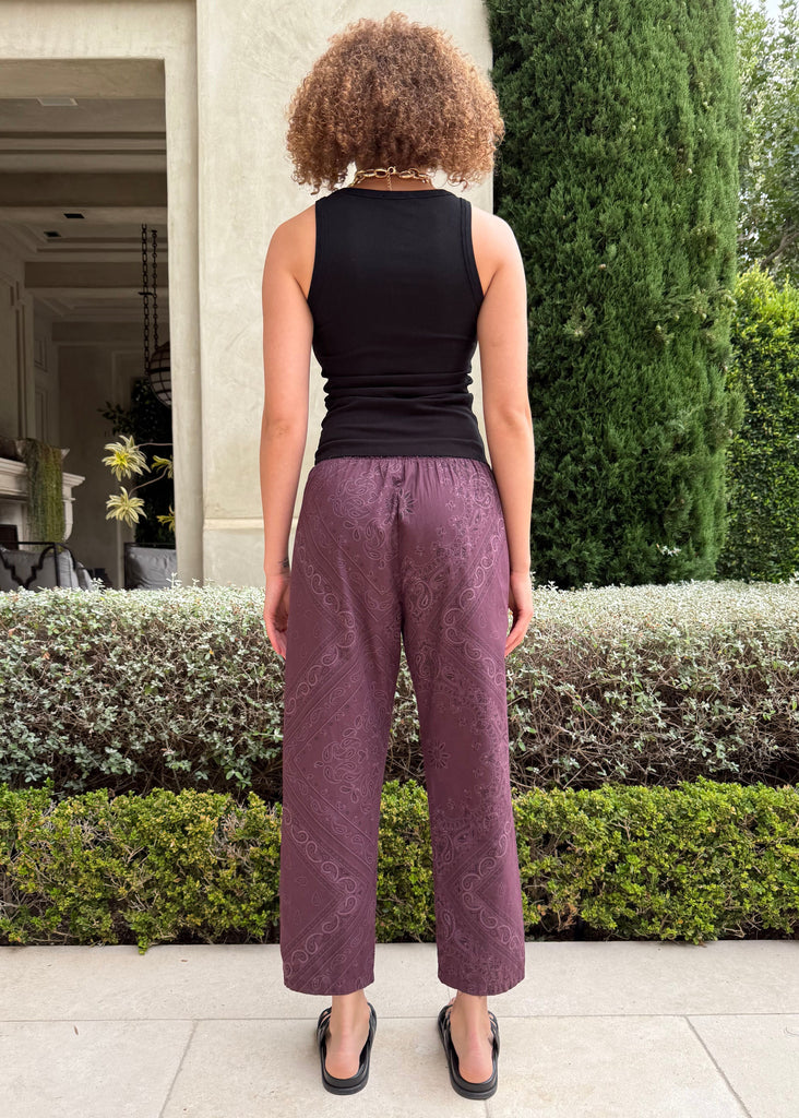 Person wearing a black sleeveless top and purple pants standing in front of a hedge.