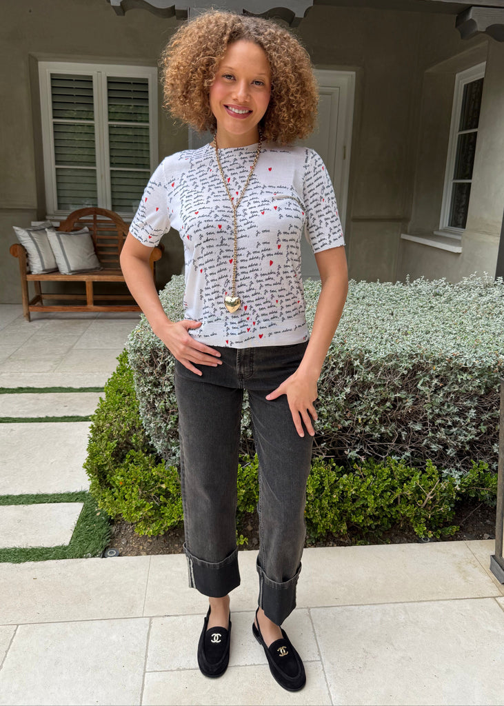 Woman standing outdoors in front of a house wearing a white t-shirt with text and dark jeans.