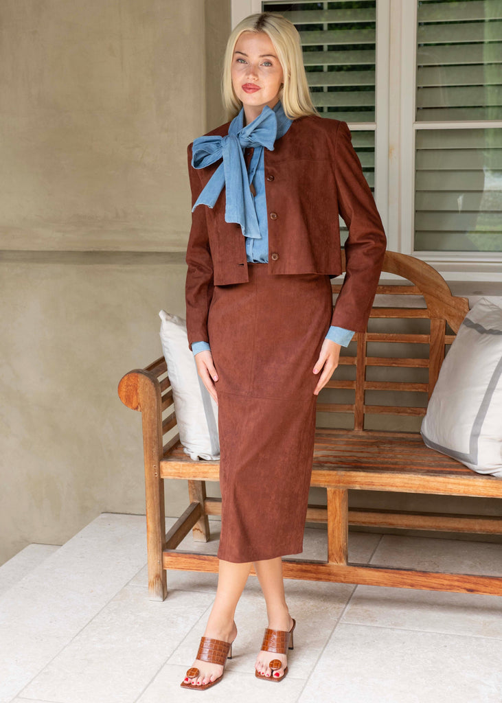 Woman in a brown outfit with a blue scarf standing in a room with a wooden bench and cushions.