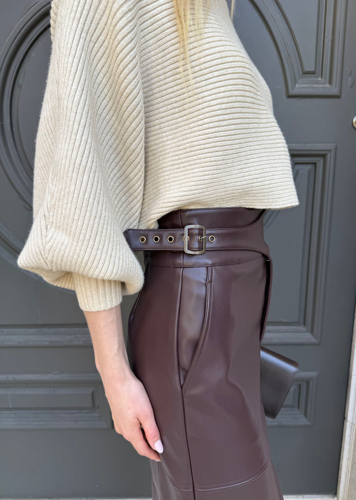 Person wearing a beige ribbed sweater and brown leather skirt in front of a gray door.