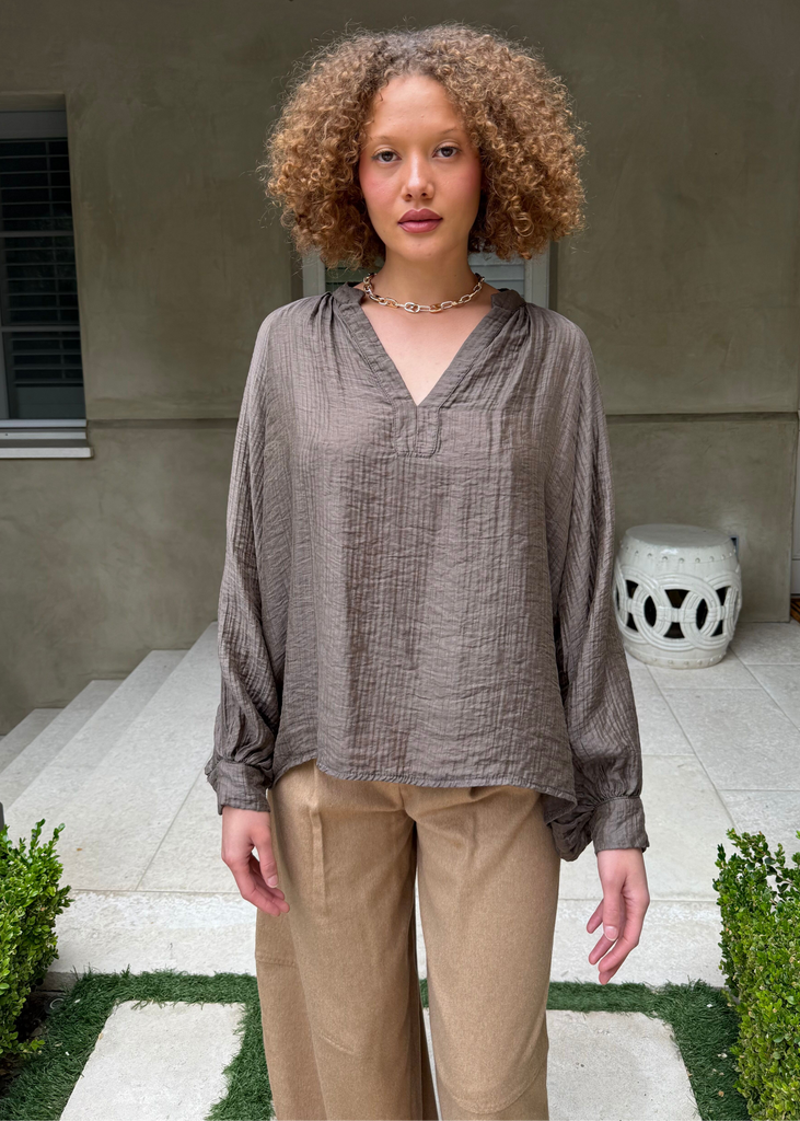 Woman standing outdoors in front of a house wearing a brown top and beige pants.