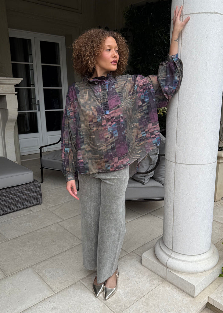 Woman in a patterned blouse and gray pants standing by a white column outdoors.