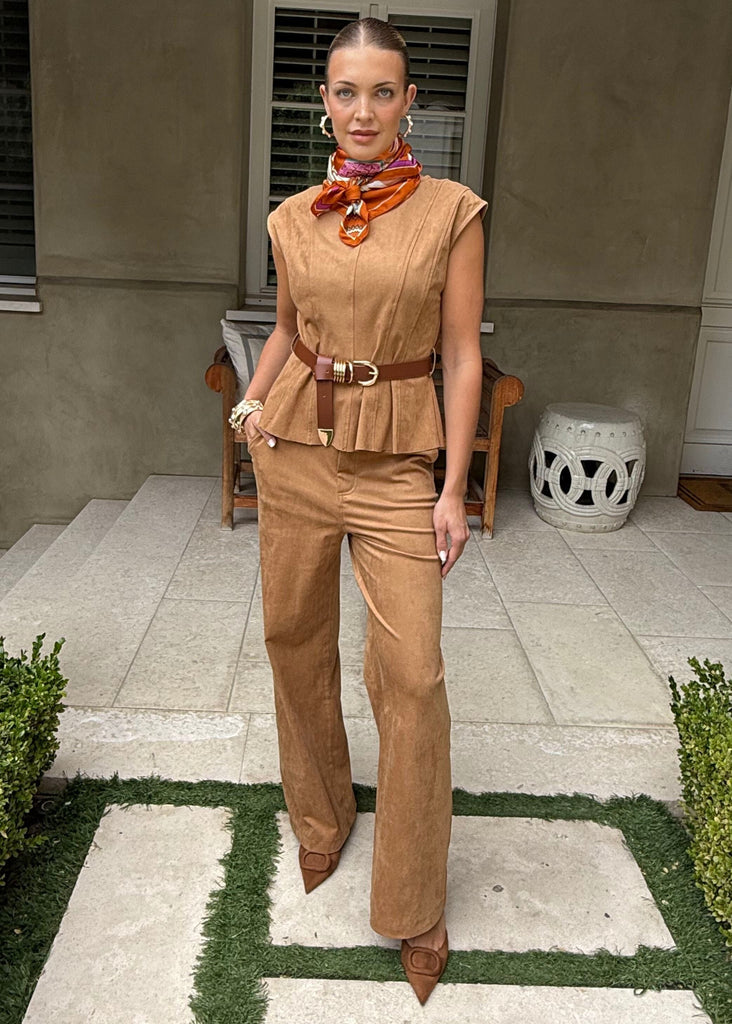 Woman in a tan outfit standing on a patio with greenery and a building in the background