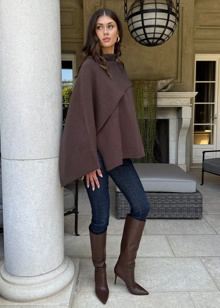Woman in a brown cape and knee-high boots standing in a stylish outdoor setting.