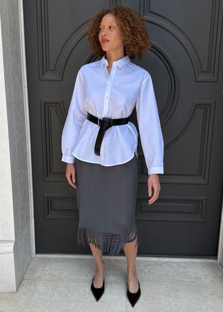 Woman wearing a white shirt, black belt, and gray skirt standing in front of a dark door.