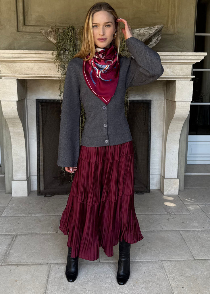 Woman in a gray jacket and burgundy skirt standing in front of a fireplace.